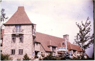 A historic photo of Eaton Hall, a stone building with a tower and a red roof, surrounded by trees.