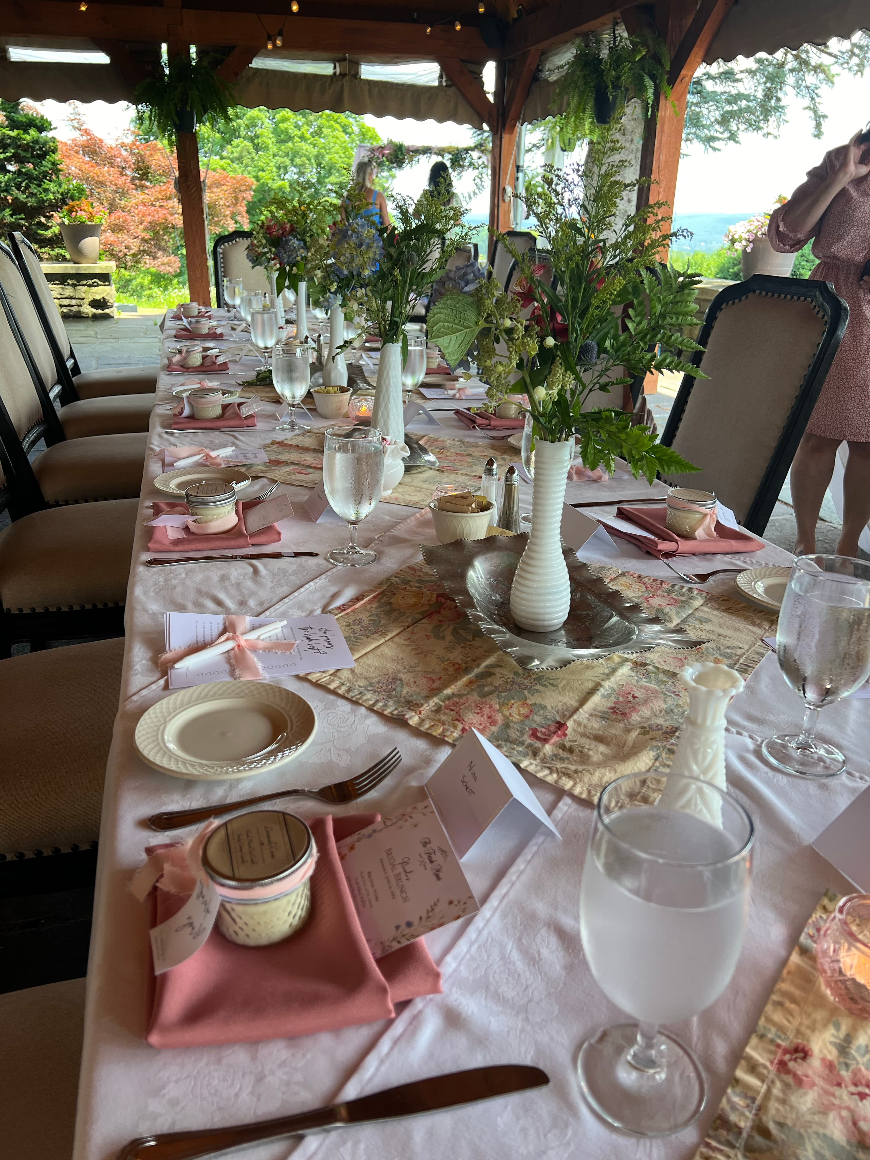 A picturesque outdoor dining area with a long table decorated for a special event, featuring floral arrangements, place settings, and a stunning view.