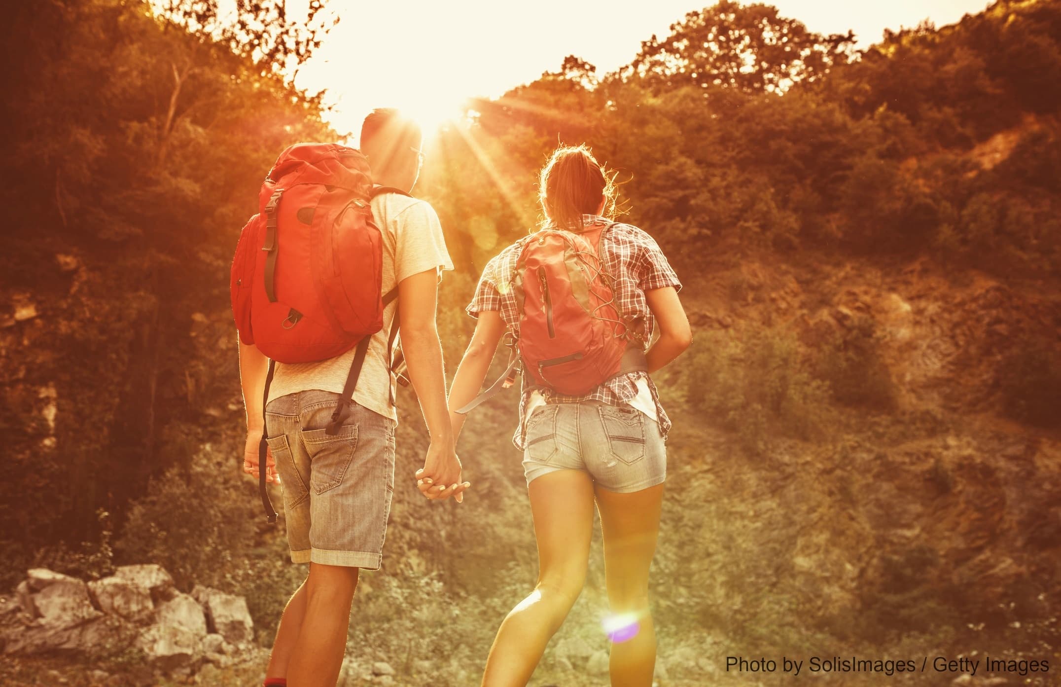A couple with backpacks walks hand in hand towards a sunset on a hiking trail.