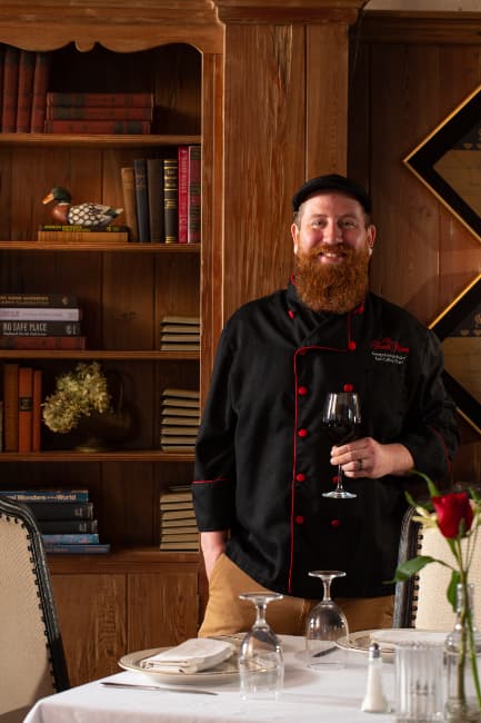 A smiling, bearded chef in a black hat and chef's coat with red trim, in a dining room in front of a table holding a glass of red wine.