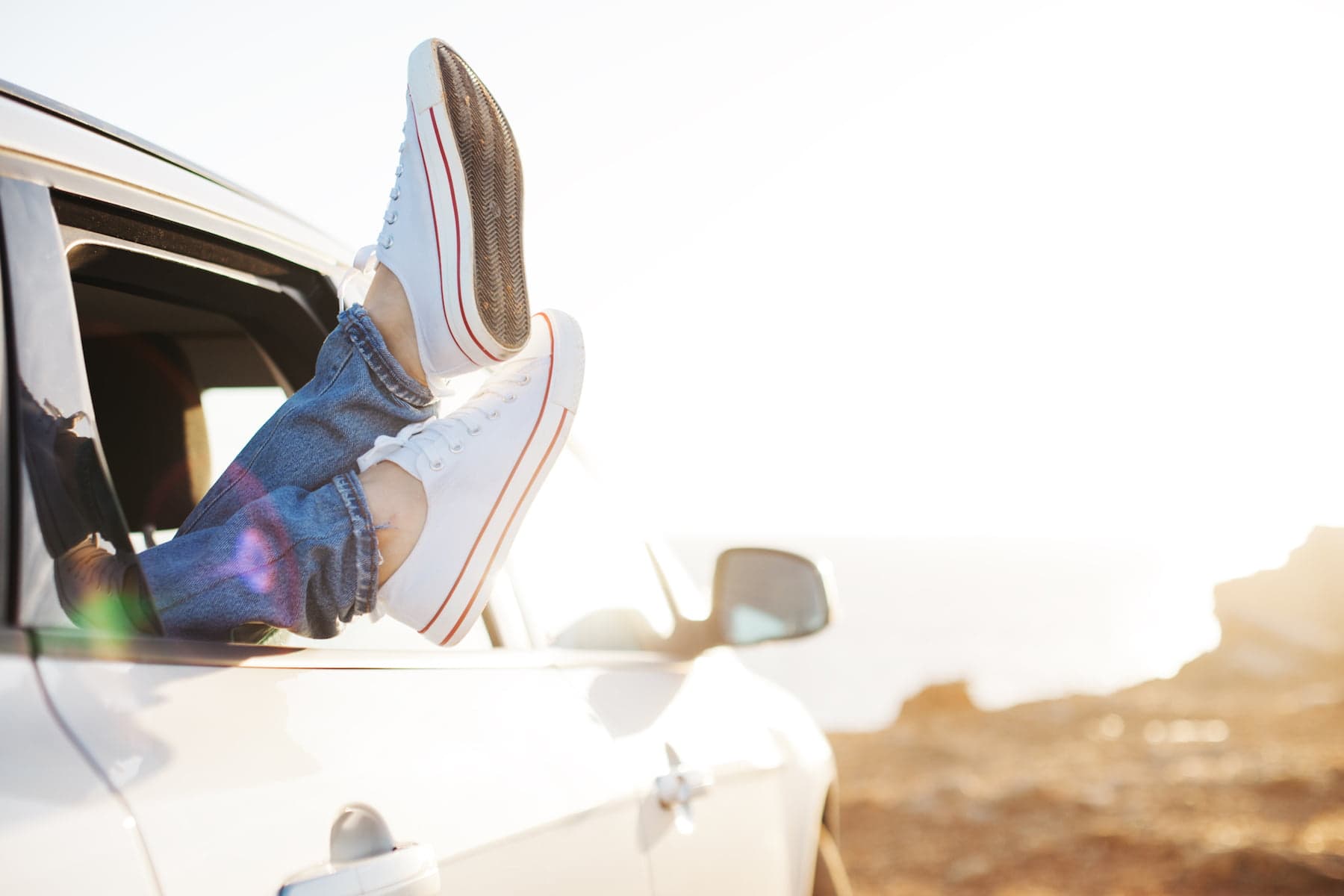 A pair of feet in white sneakers rests on the doorframe of a car with the ocean in the background.
