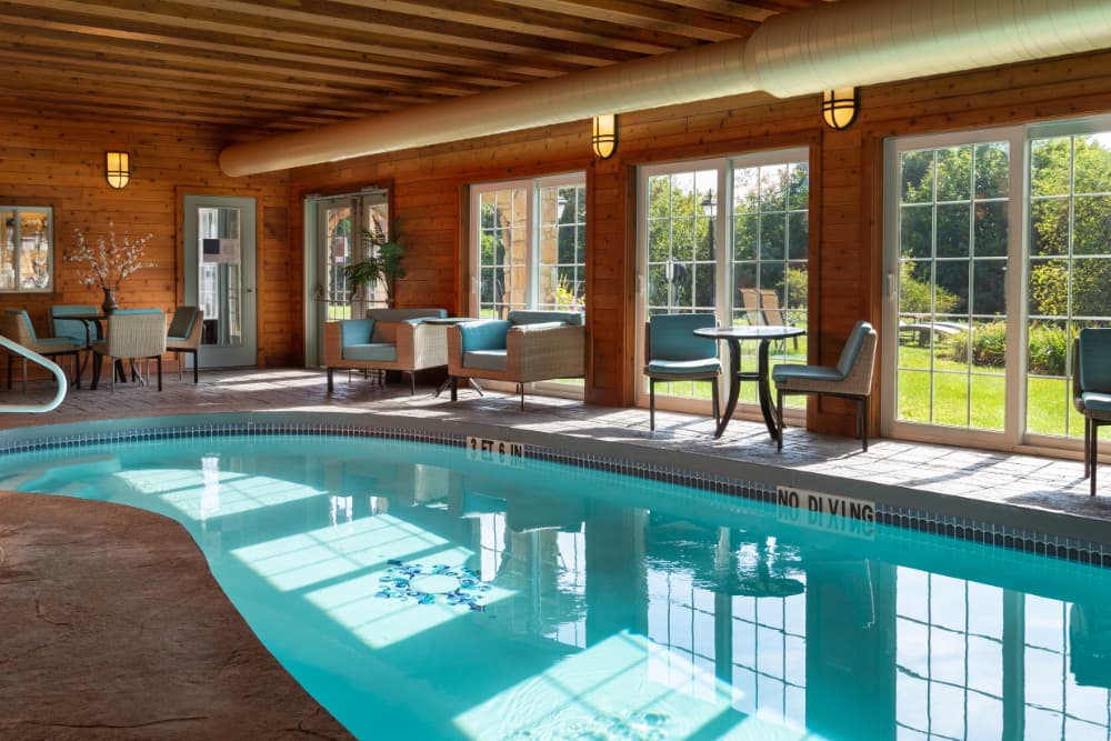 An indoor swimming pool with large windows overlooking a lush green lawn. The pool is surrounded by comfortable seating areas with armchairs and a small table. The ceiling is high with exposed wooden beams.