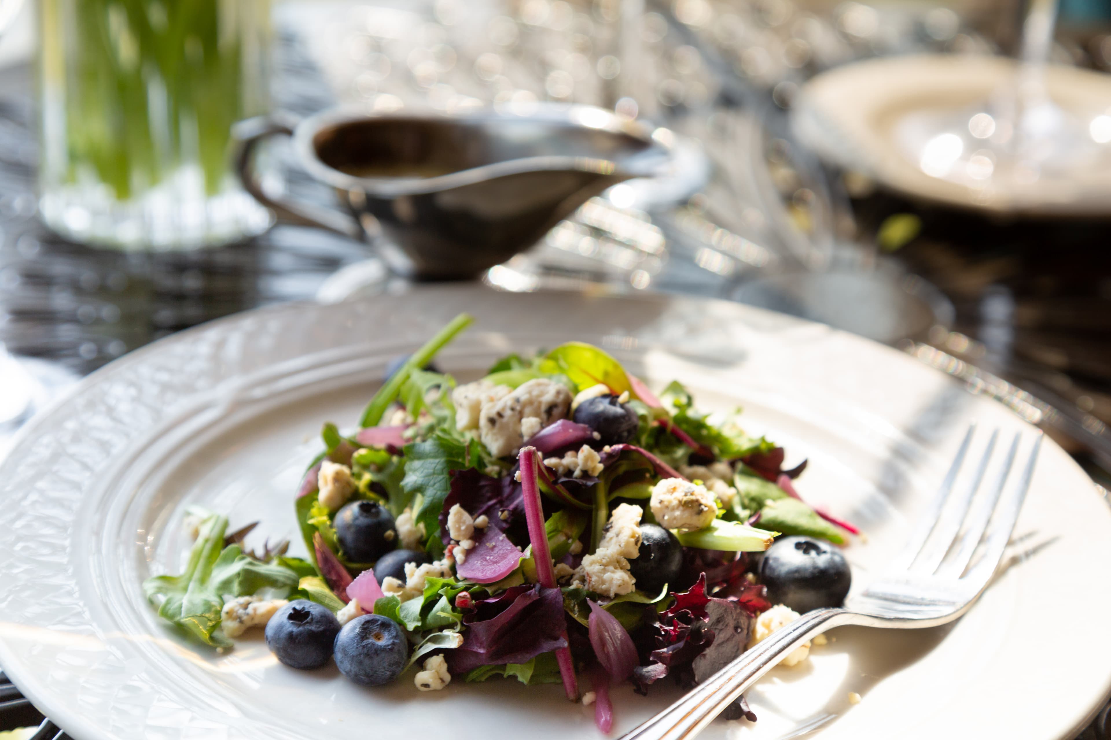 A fresh salad with mixed greens, blueberries, crumbled blue cheese, and a balsamic vinaigrette dressing. The salad is served on a white plate with a fork and a small gravy boat filled with dressing in the background.