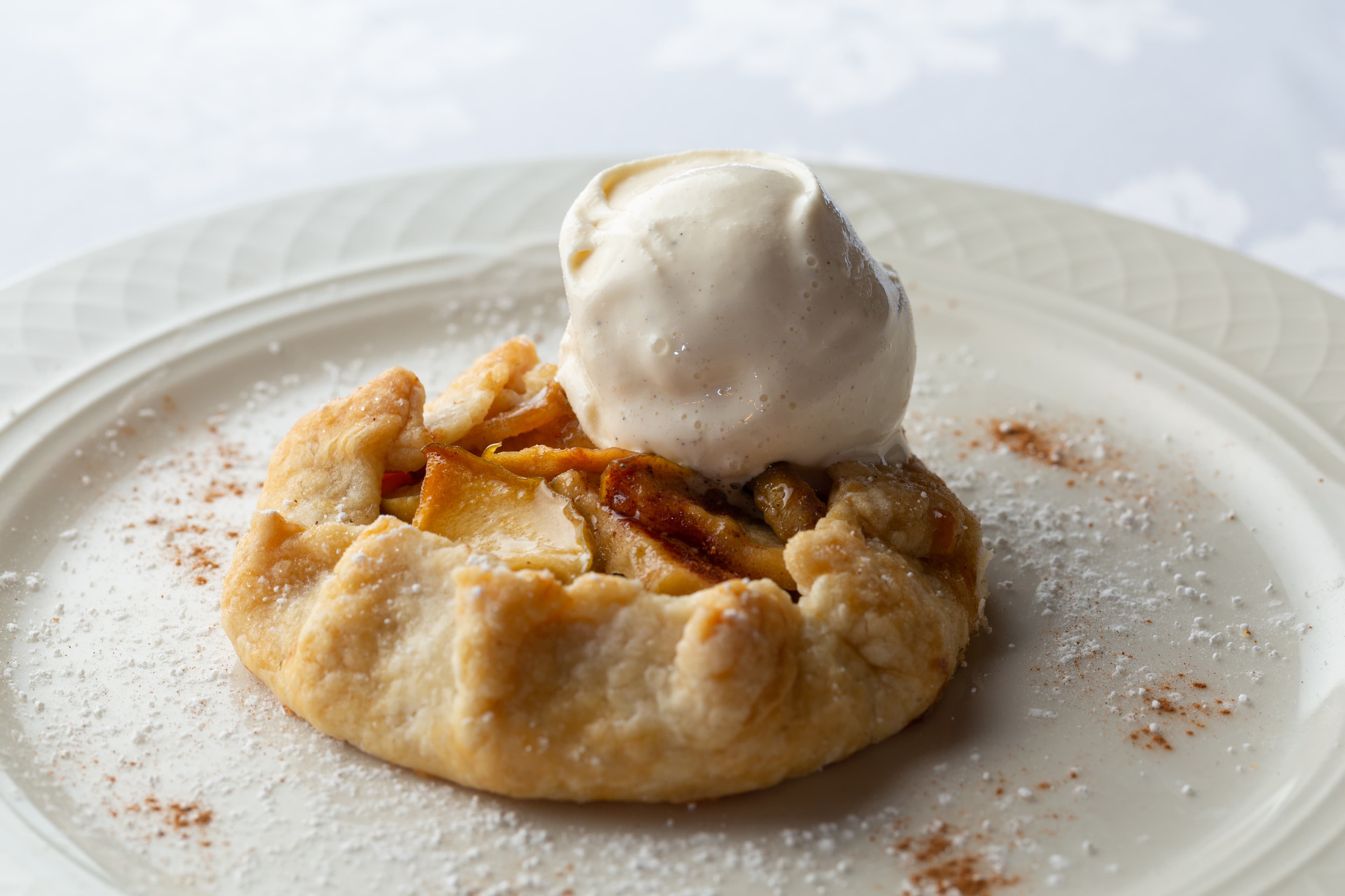 A delicious apple galette served with a scoop of vanilla ice cream. The galette is a free-form pie with a flaky crust and a sweet apple filling. It is dusted with powdered sugar and cinnamon.