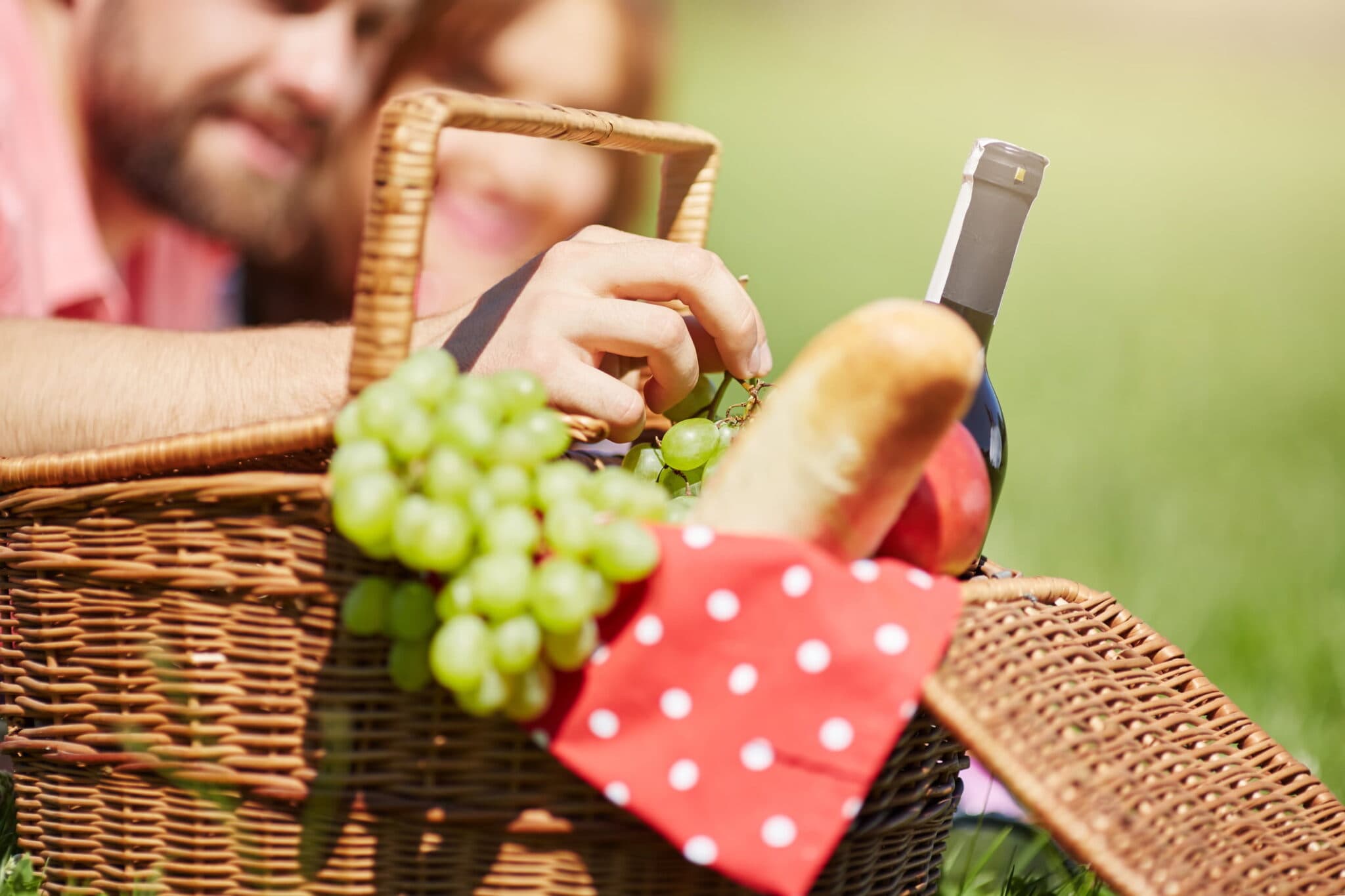 A picnic basket with grapes, a loaf of bread, and a bottle of wine, with two people partially visible in the background.