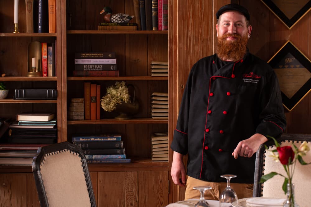 A smiling, bearded chef in a black hat and chef's coat with red trim, in a room with bookshelves behind him, in front of a table set for an elegant dinner.