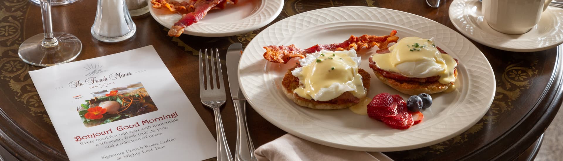 A delicious breakfast spread on a table in front of a fireplace. The meal includes French toast, eggs Benedict, fruit salad, orange juice, coffee, and bacon. The table is set with a white tablecloth, silverware, and a menu with the words "Bon Jour! Good Morning!" written on it.