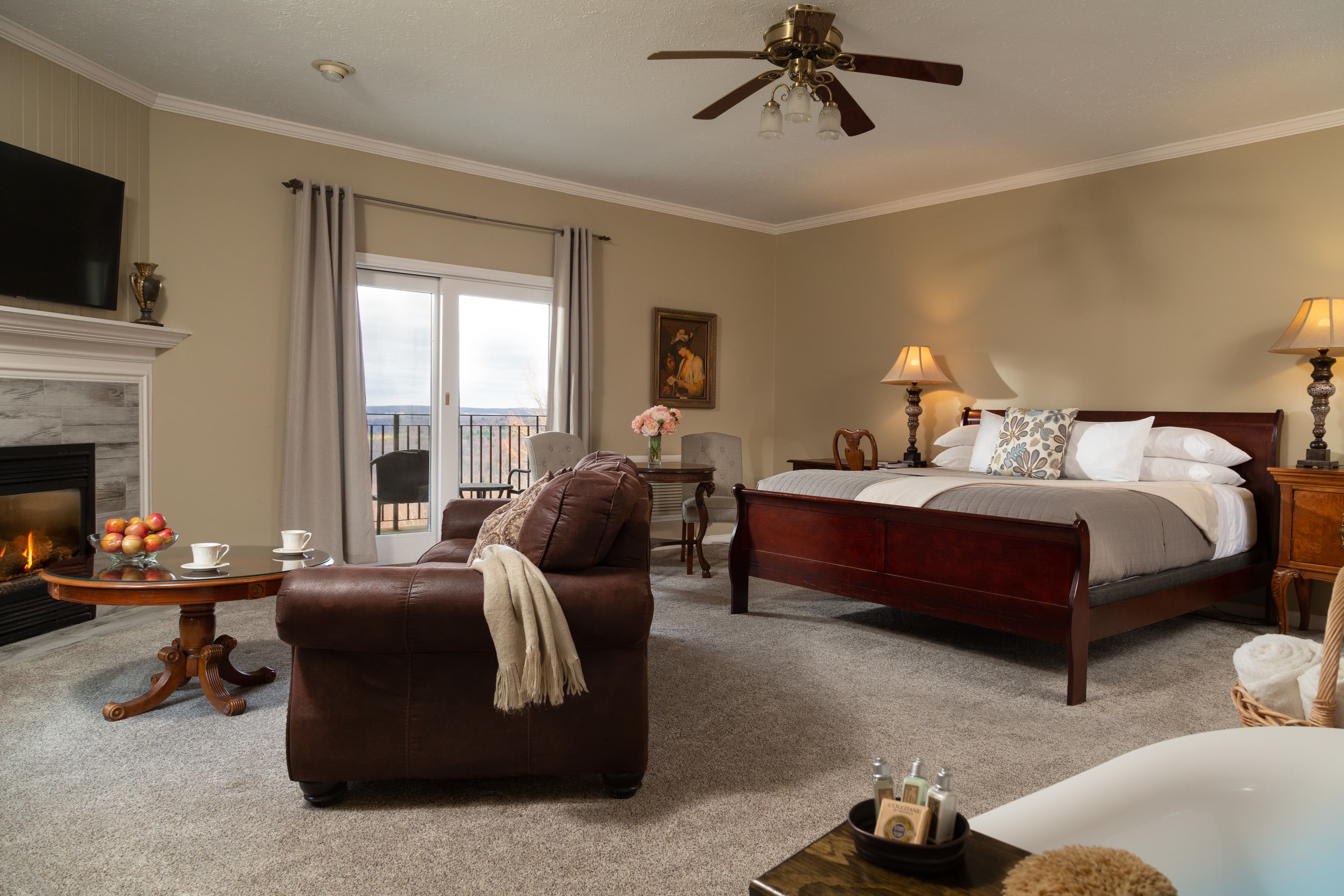 A cozy, well-decorated hotel room featuring a king bed, a leather armchair, a fireplace, and a scenic view through large windows.