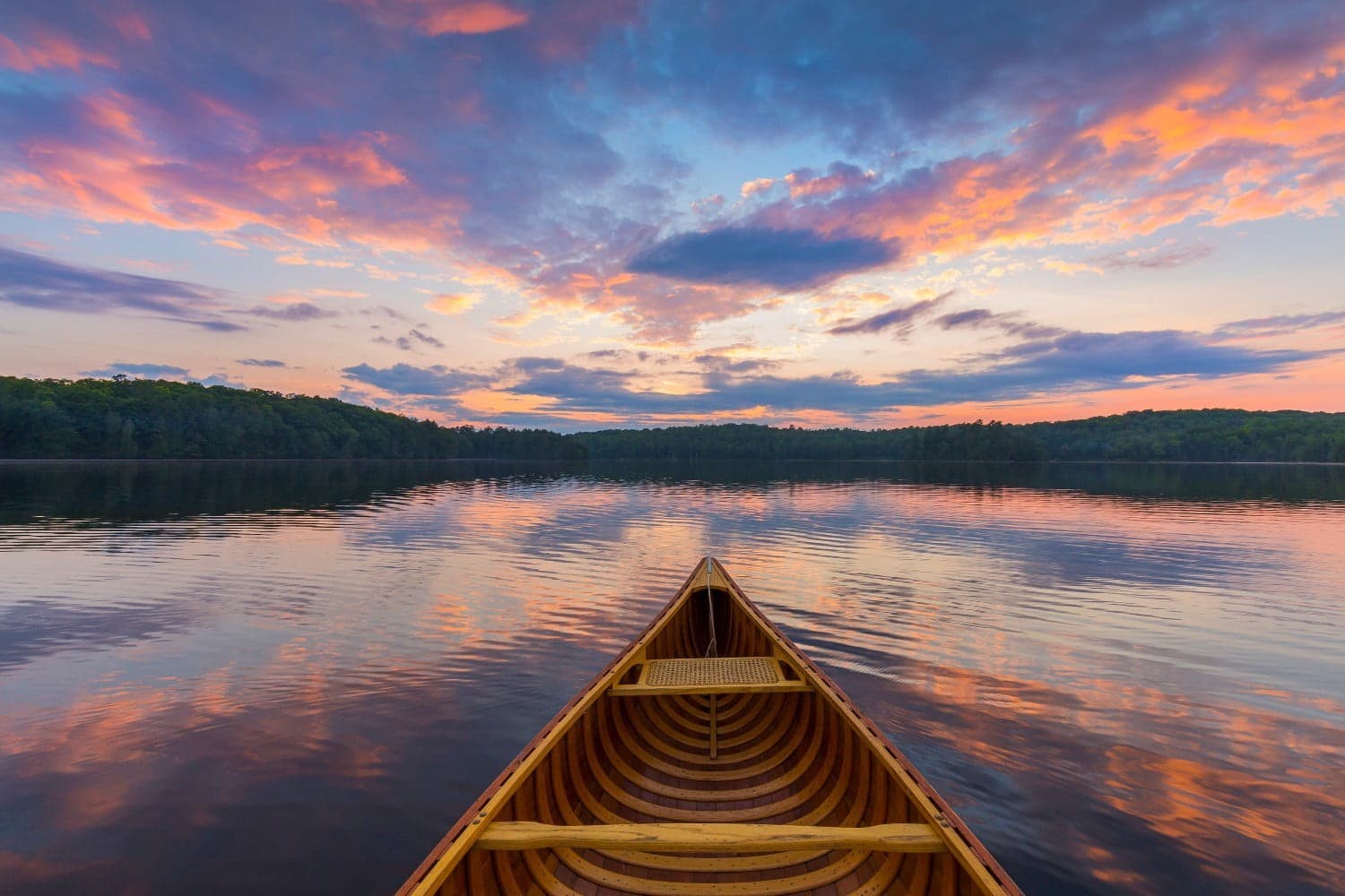 A wooden canoe floats on a calm lake reflecting a colorful sunset.