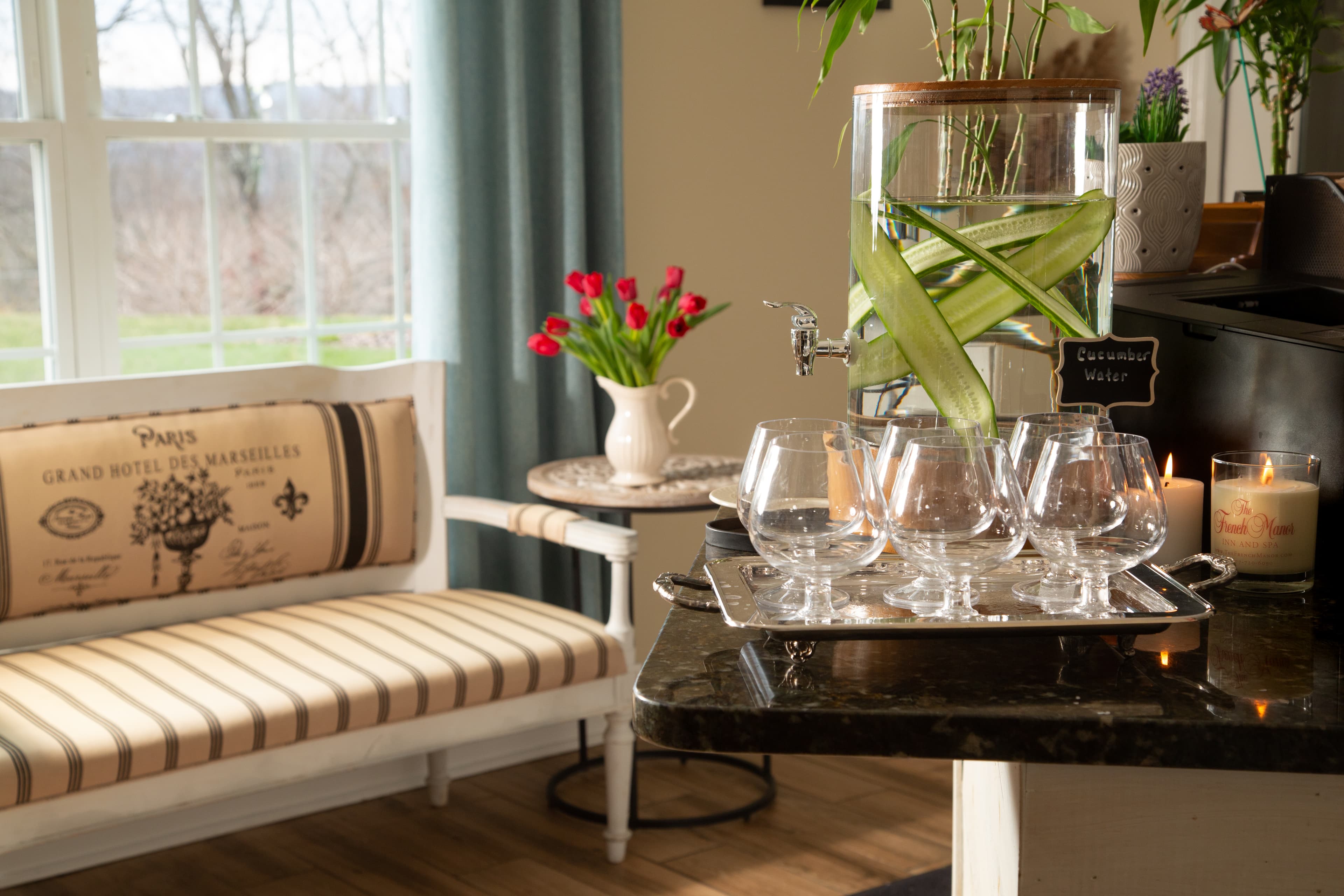 A cozy sitting area with a striped bench, a vase of tulips, and a water dispenser filled with cucumber-infused water. There are also glasses and candles on a tray.