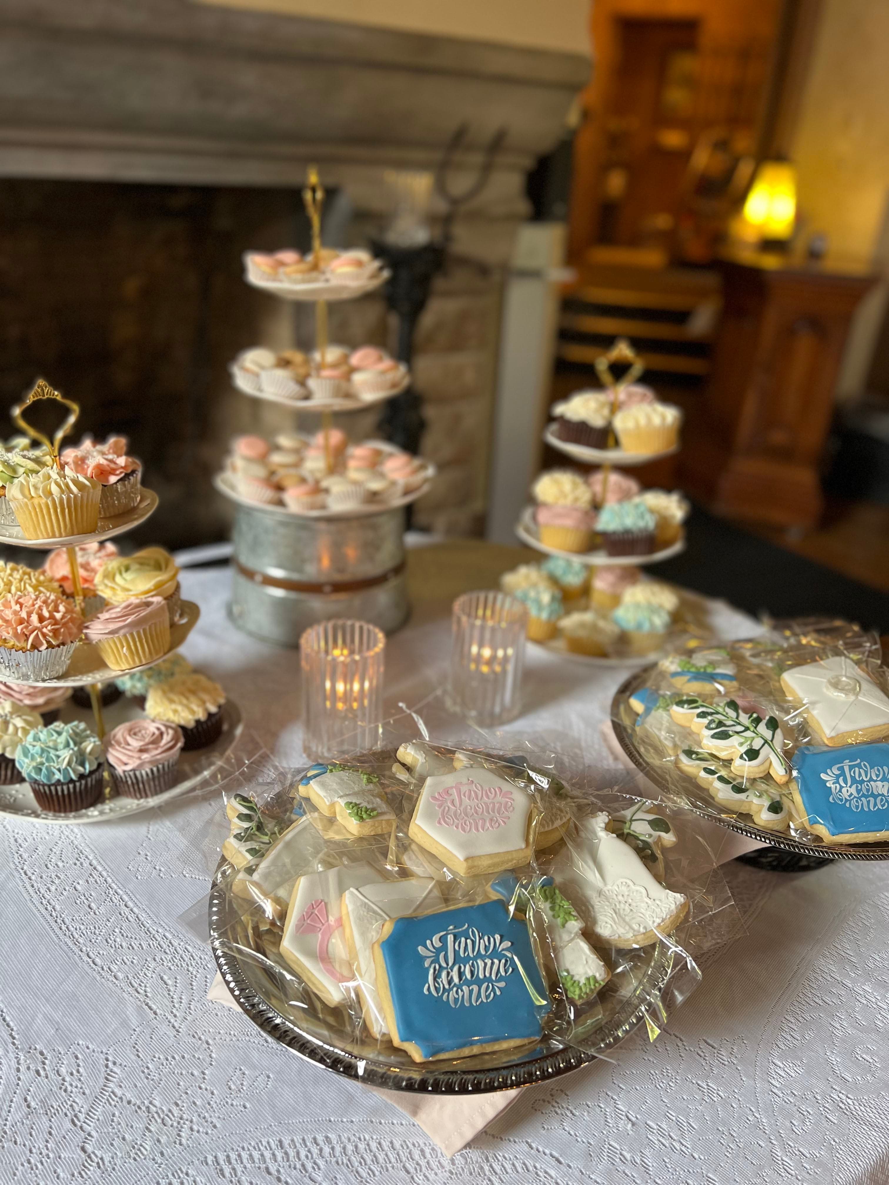 A tiered stand filled with cupcakes and a silver platter of decorated sugar cookies, with a fireplace and a lamp in the background.