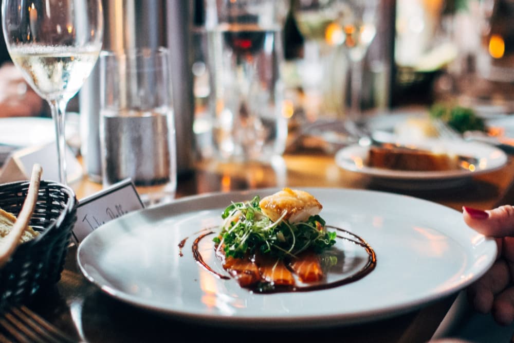 A plate of salmon with a sauce and greens, served with a glass of white wine with more plates of the same on the table.