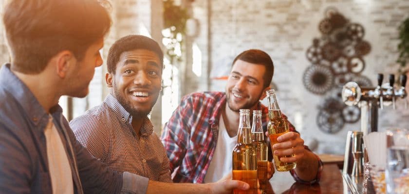 Three friends sitting at a bar, smiling and holding bottles of beer.