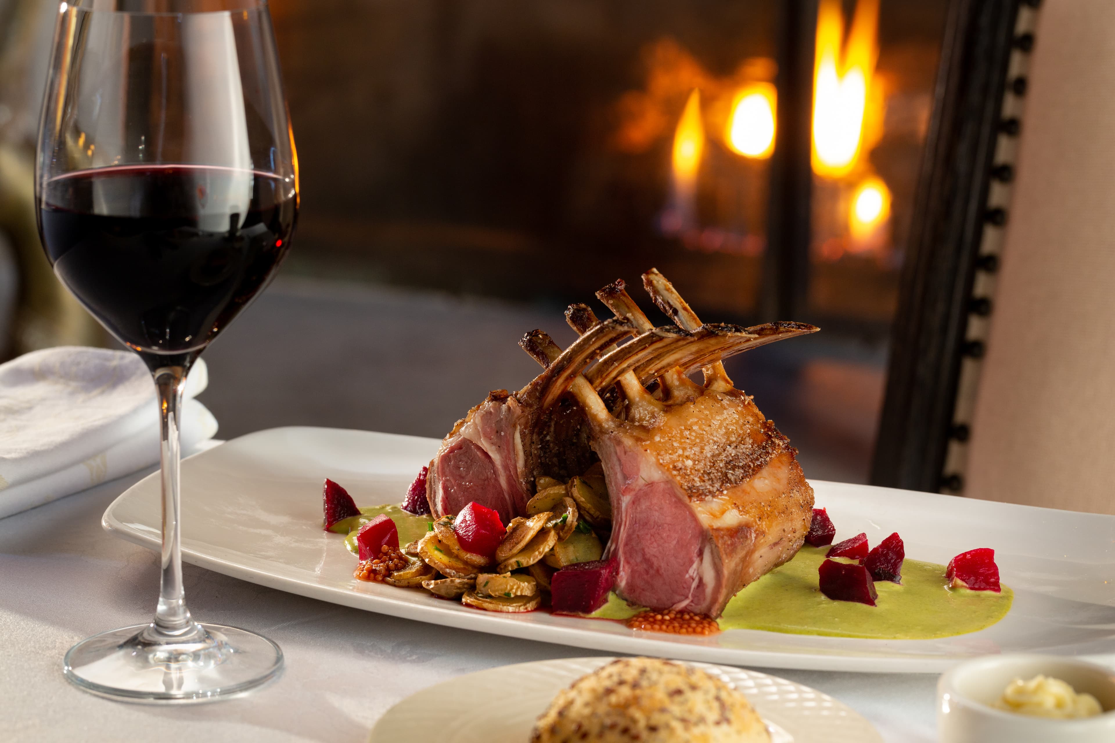 A plate 2 elegantly presented rack of lambs served with a green sauce, mushrooms, and roasted beets. A glass of red wine is placed next to the plate, and a fireplace is visible in the background.