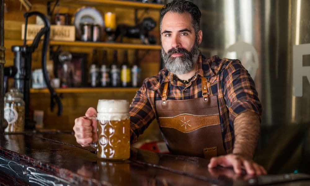 A bartender holding a glass of beer at a brewery bar, with brewing equipment and shelves in the background.