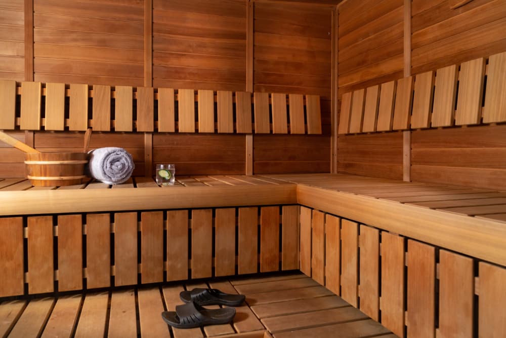 A wooden sauna with benches, a bucket, a towel, a glass of water., and a a pair of black sandals on the floor. The walls and ceiling are lined with wood panels.