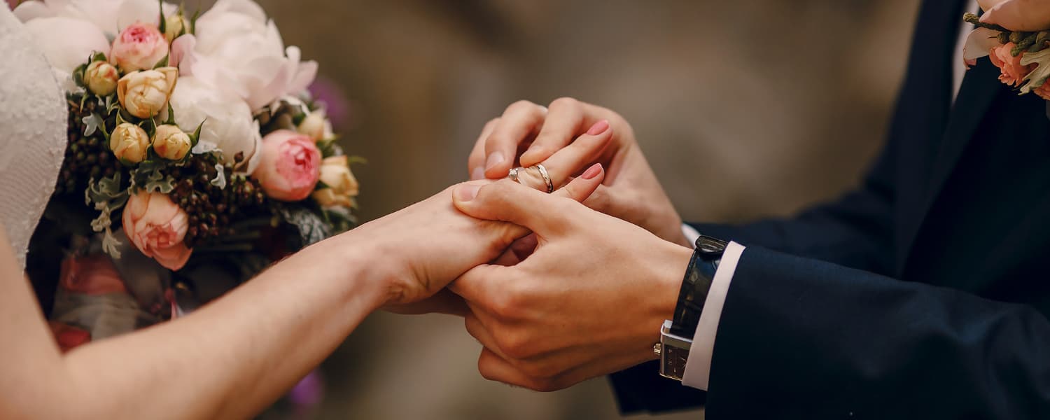 A bride and groom dressed in wedding attire with the woman holding a bouquet of pink and yellow flowers, and the room putting a wedding ring on the woman's ring finger.