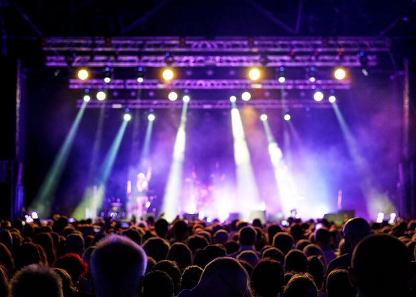 A crowd of concertgoers watches a performance on a vibrant stage illuminated by colorful lights.