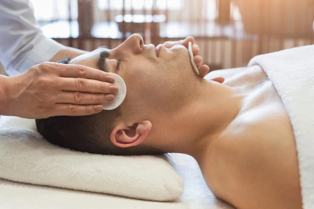 A man laying on his back, face up, on a massage table with a thin pillow under his head, getting a facial treatment.
