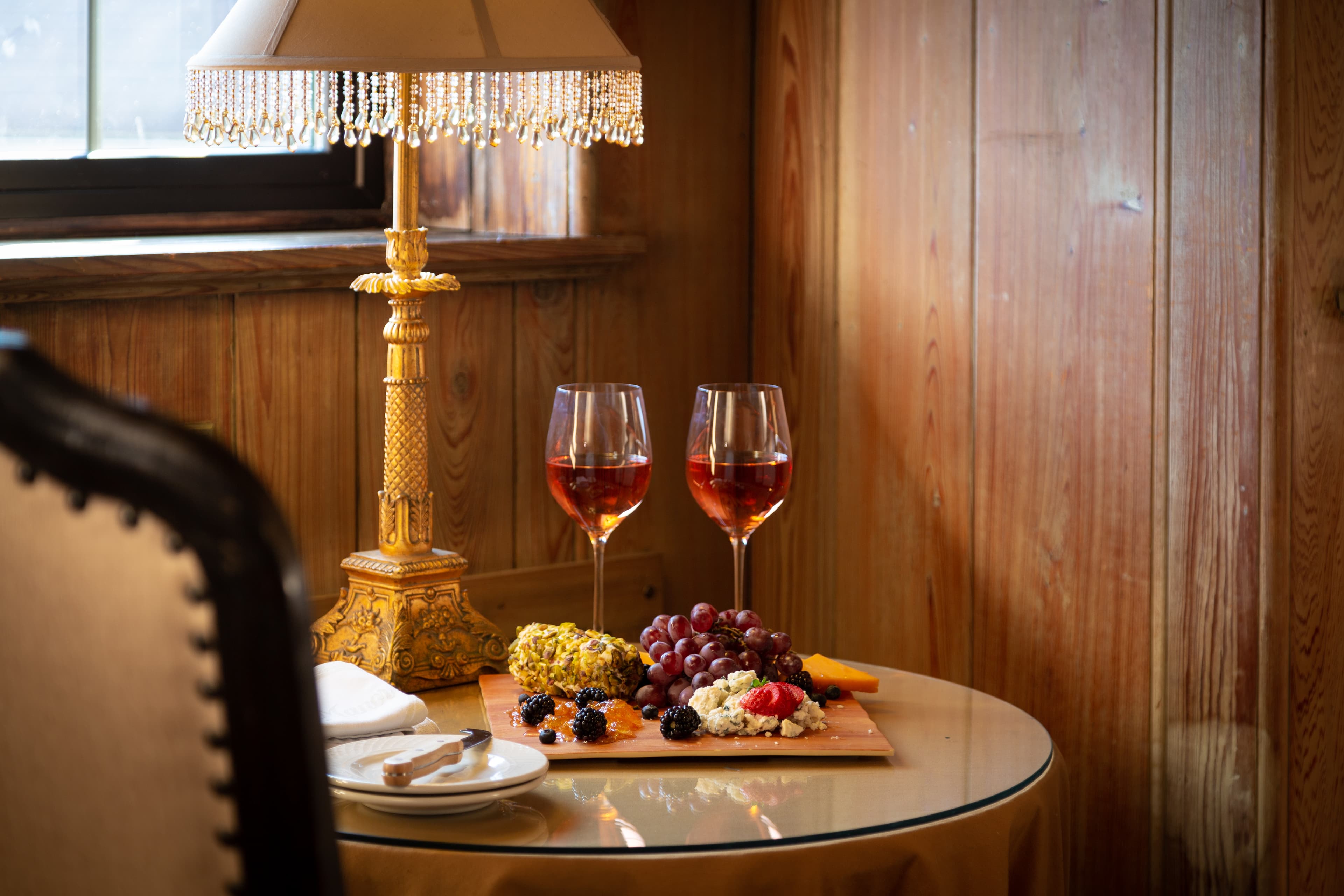 A cozy setting with a lamp, two glasses of rosé wine, and a charcuterie board on a glass table.