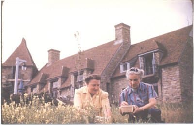 A historic photo of Eaton Hall, a stone building with a tower and a red roof, surrounded by trees, with 2 women in the foreground picking berries.