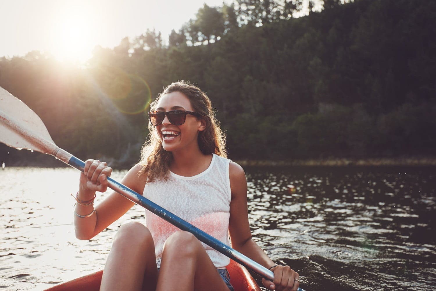 A woman with curly hair and sunglasses smiles while kayaking on a calm lake.