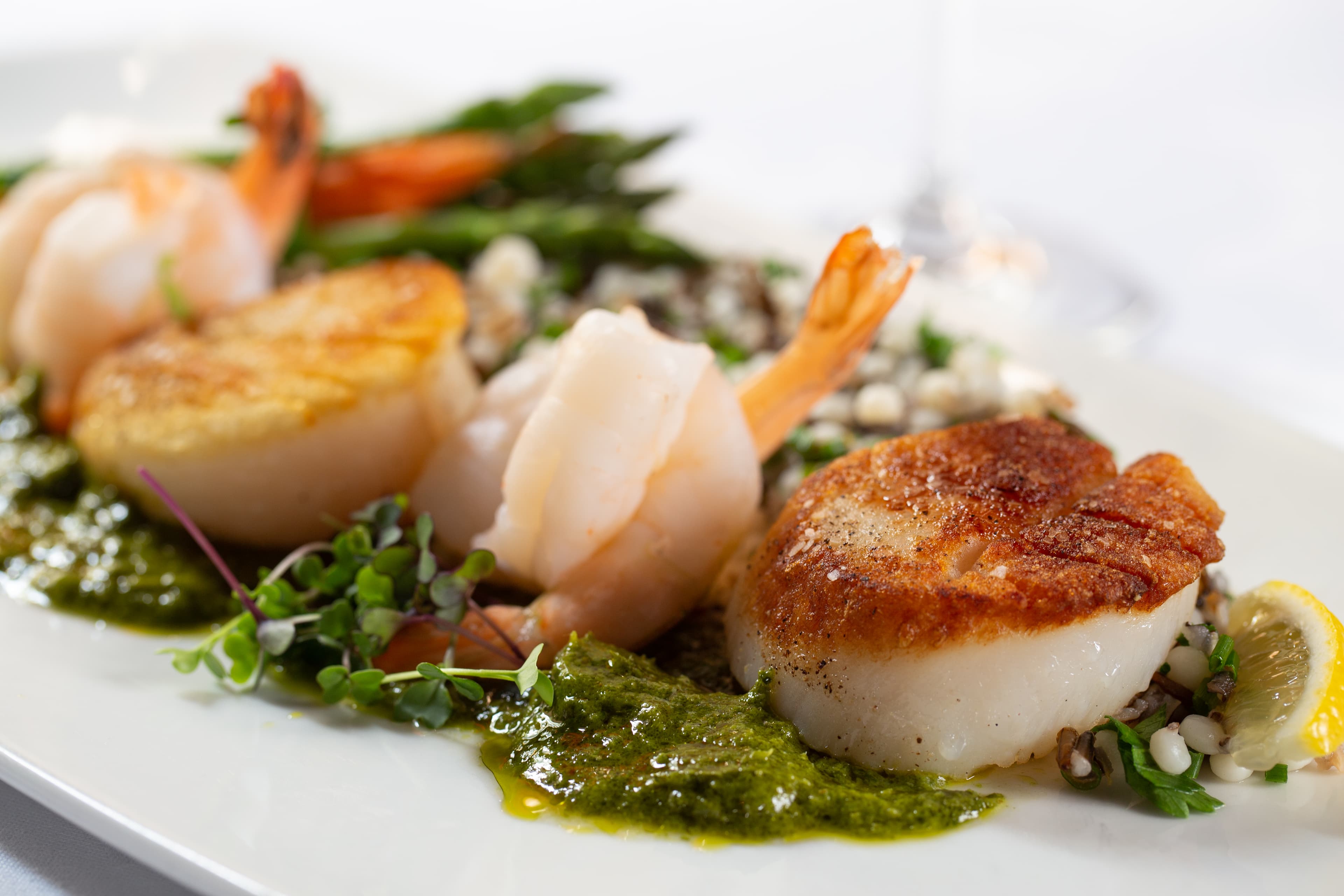 A plate of seared scallops and shrimp served with a bed of wild rice and asparagus. A green pesto sauce is drizzled over the dish, and a lemon wedge is placed on the side.