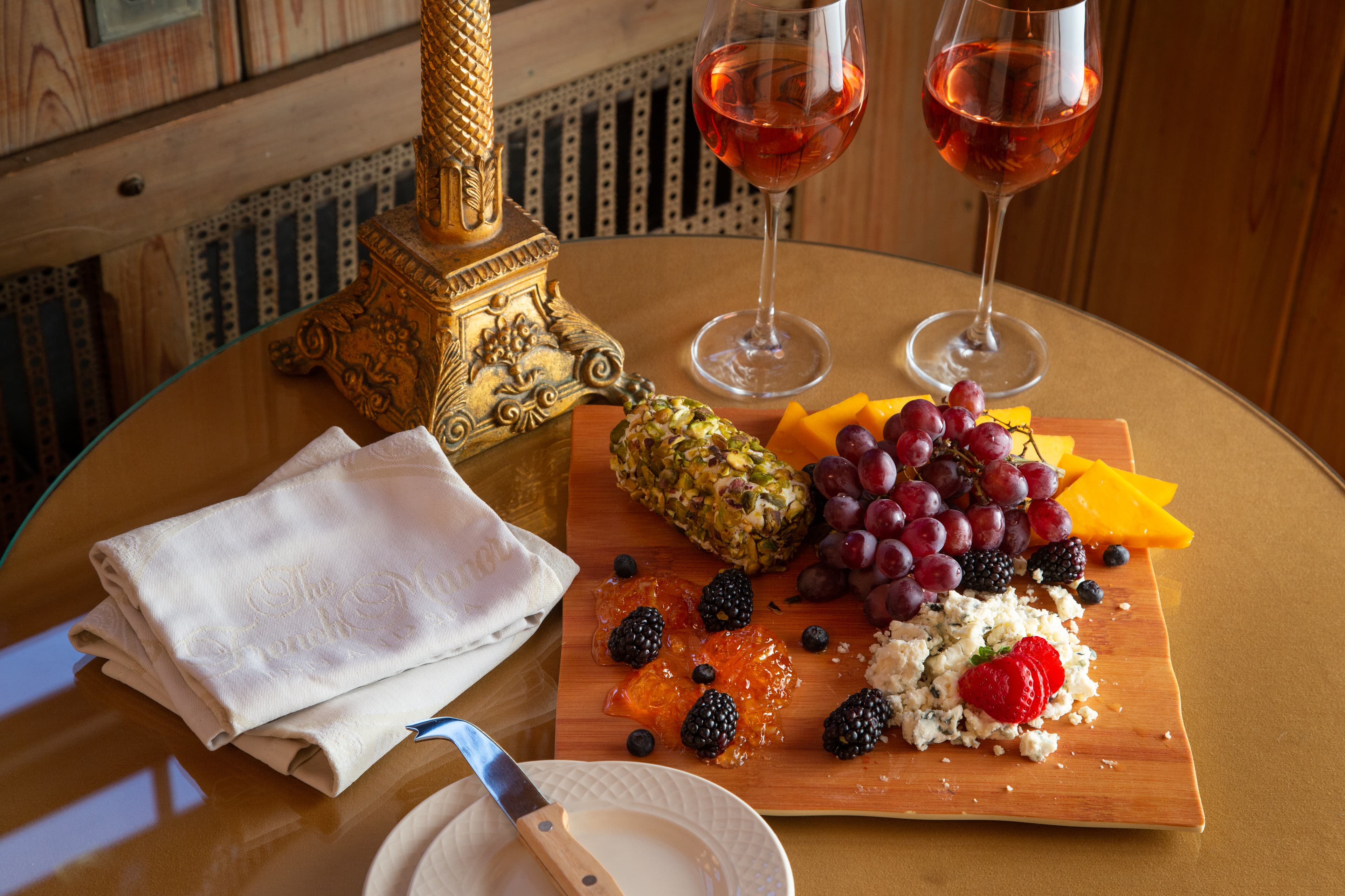 A charcuterie board with a variety of cheeses, fruits, and crackers. There are two glasses of rosé wine, a cheese knife, and napkins on the table. A gold candlestick is placed next to the board.