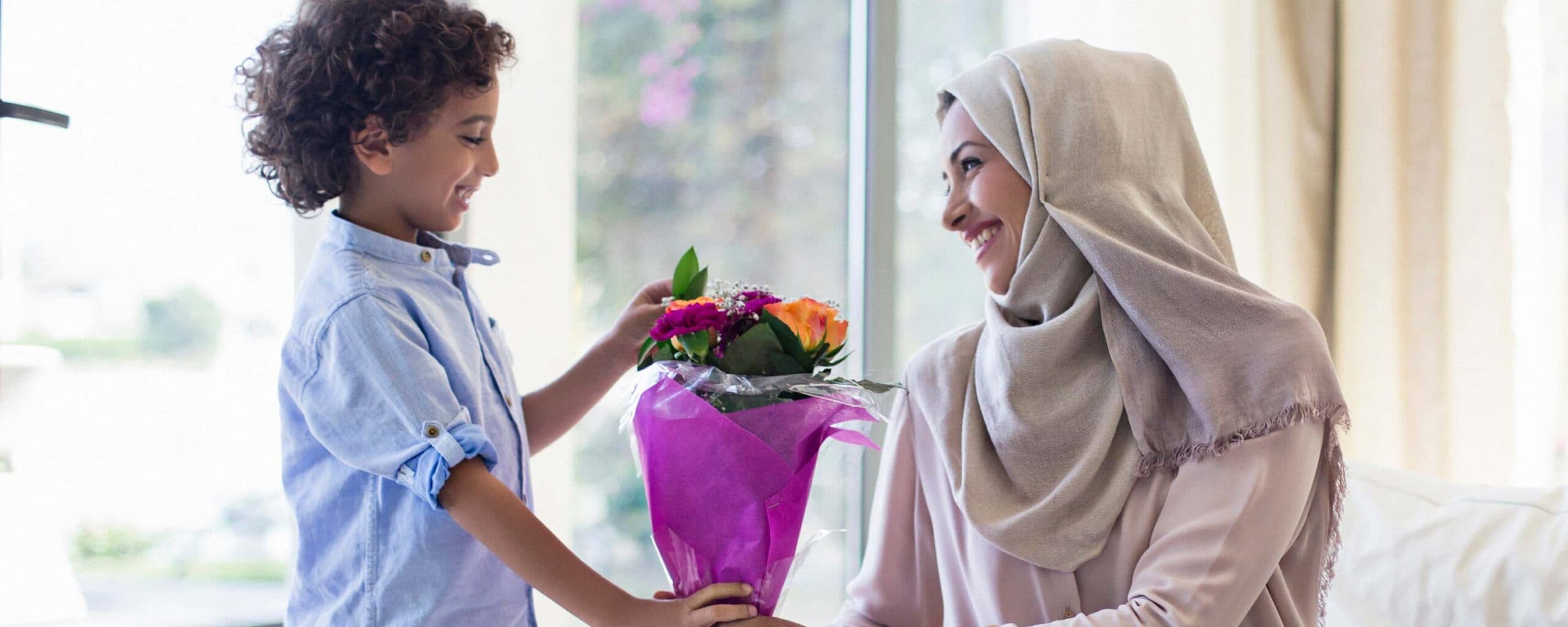 A young boy hands a bouquet of flowers to a smiling woman wearing a hijab.