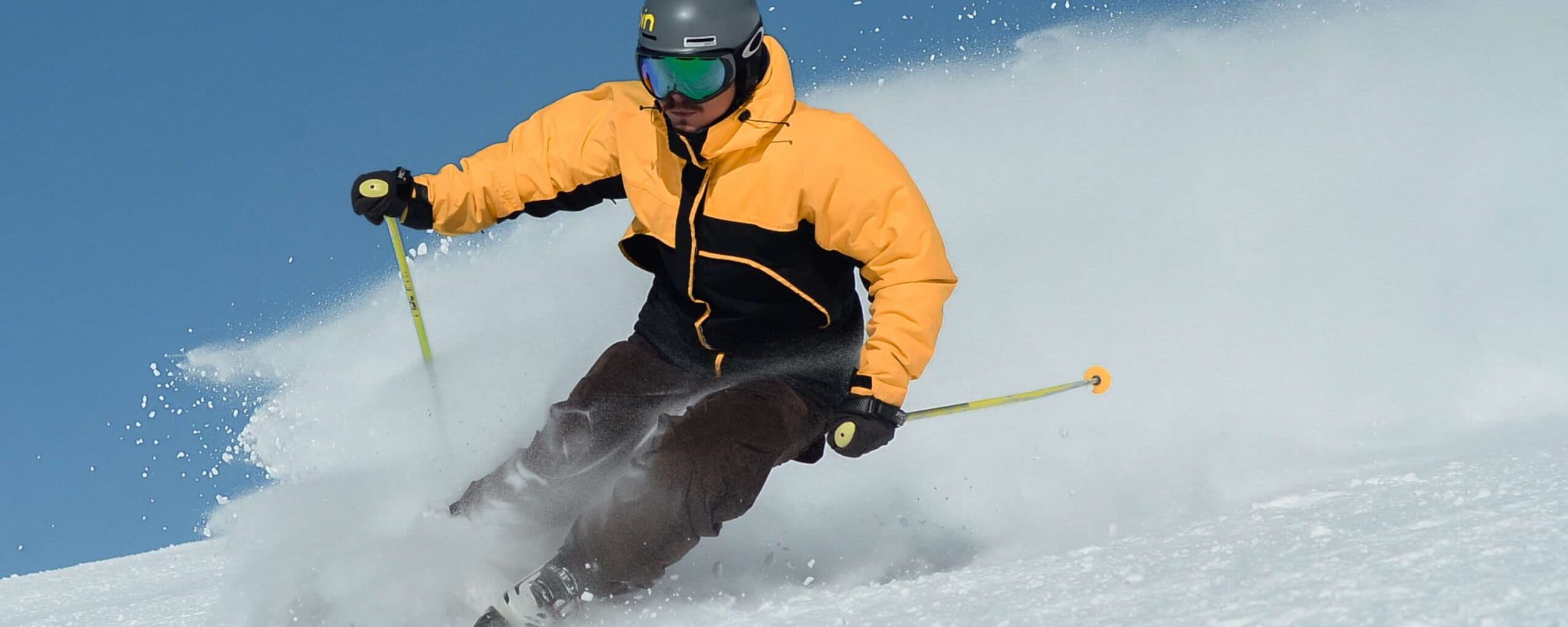 A skier in a yellow and black jacket carves through fresh snow against a clear blue sky.