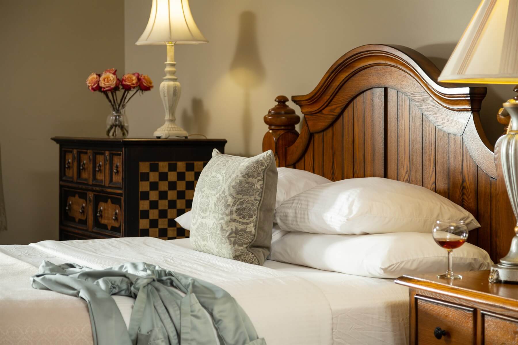 A neatly made bed with decorative pillows, a glass of wine, and a vase of flowers in a cozy bedroom setting.