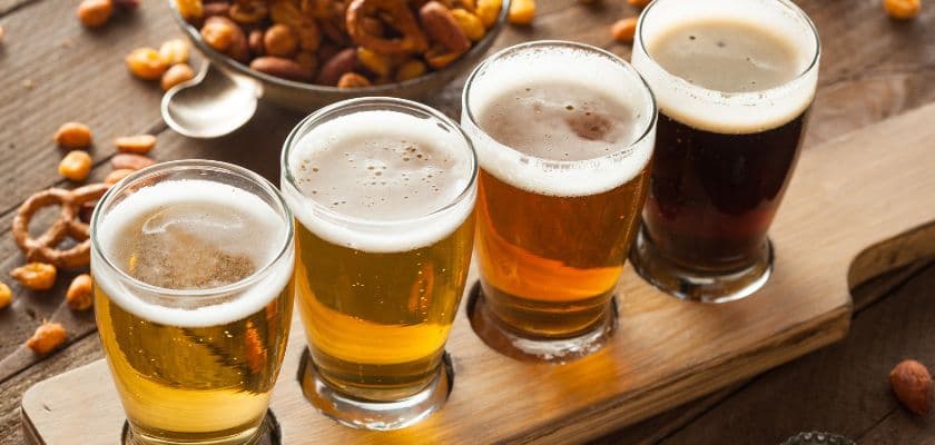 A beer flight with four glasses of craft beer on a wooden board, with pretzels and snacks nearby.