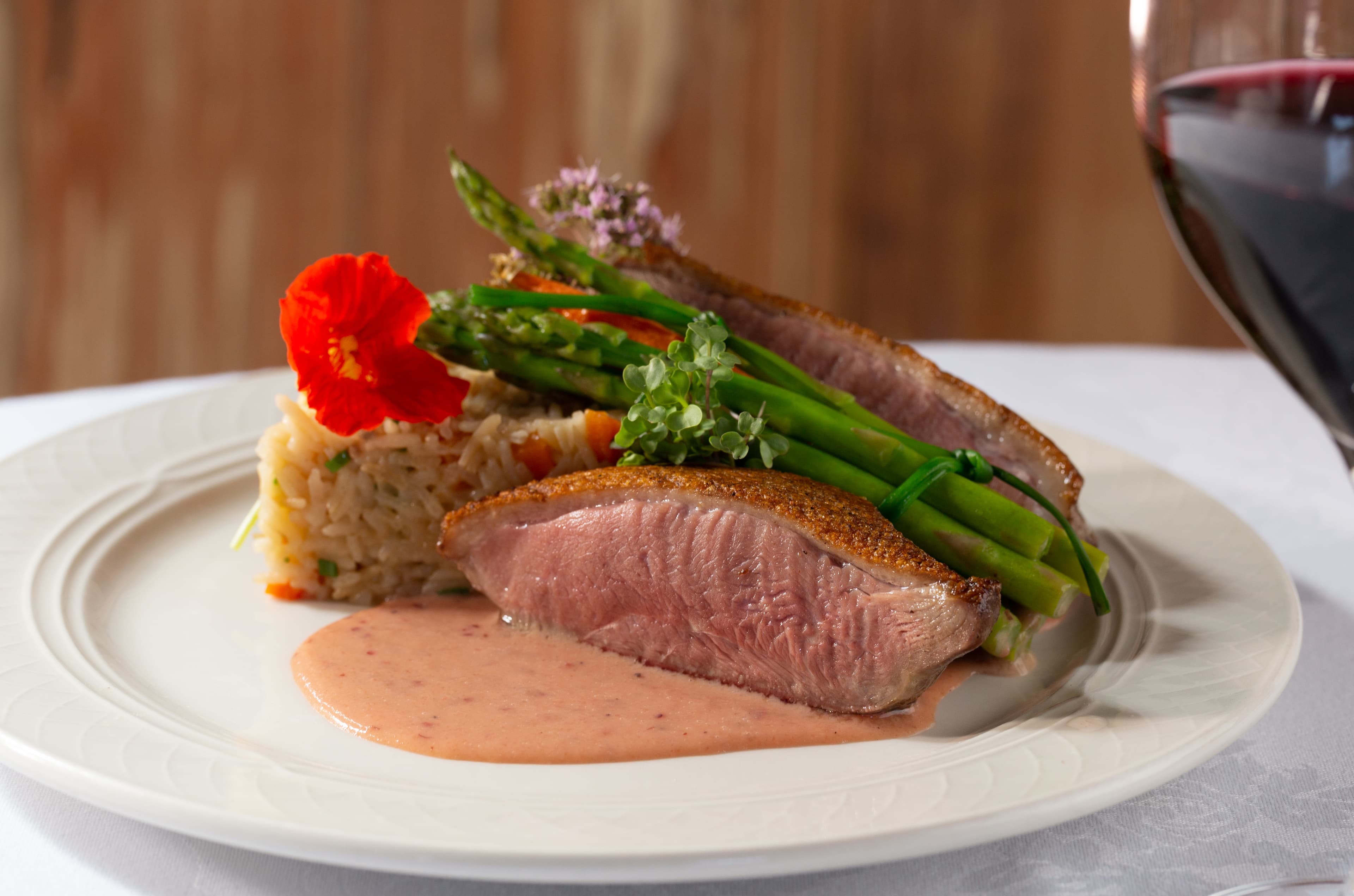 A plate of seared duck breast with a pink sauce, served with a bed of rice pilaf and asparagus. A flower is placed on the plate for garnish, and a glass of red wine is placed next to it.