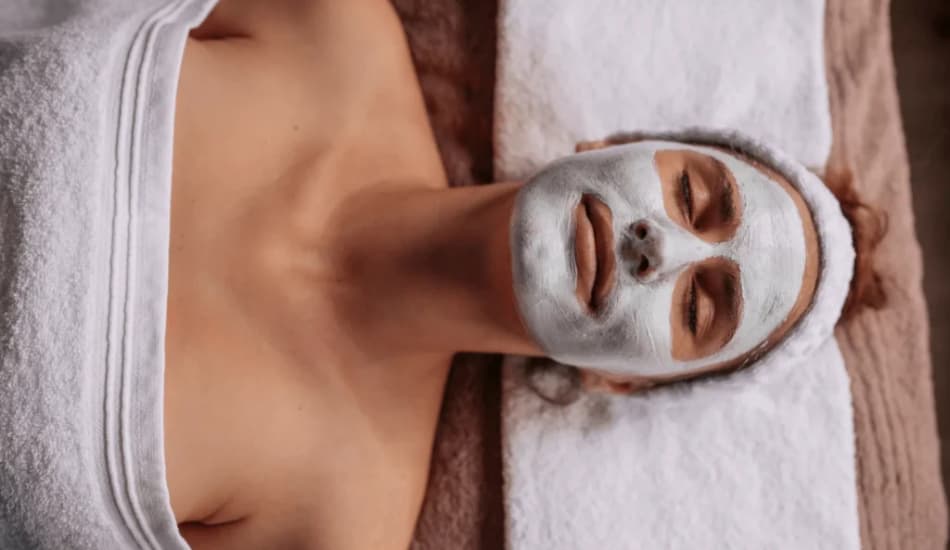 A woman lying on a table with her head on a white towel, and her face covered in a white spa mask, getting a facial