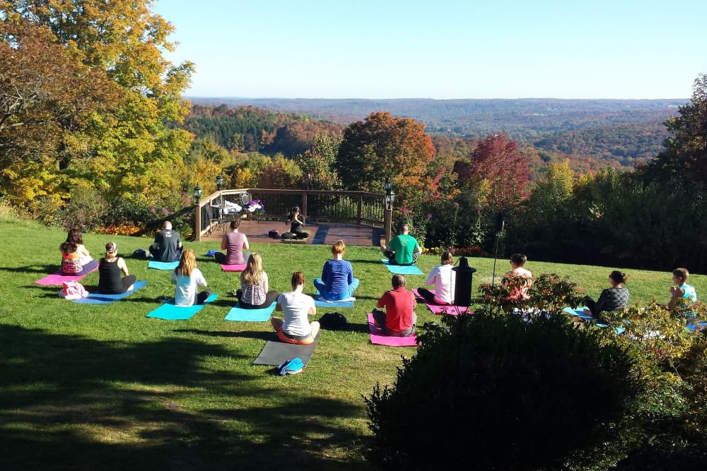 A group of people practicing yoga outdoors on a grassy hill with a scenic view of trees with fall foliage and the Poconos mountains in the background.