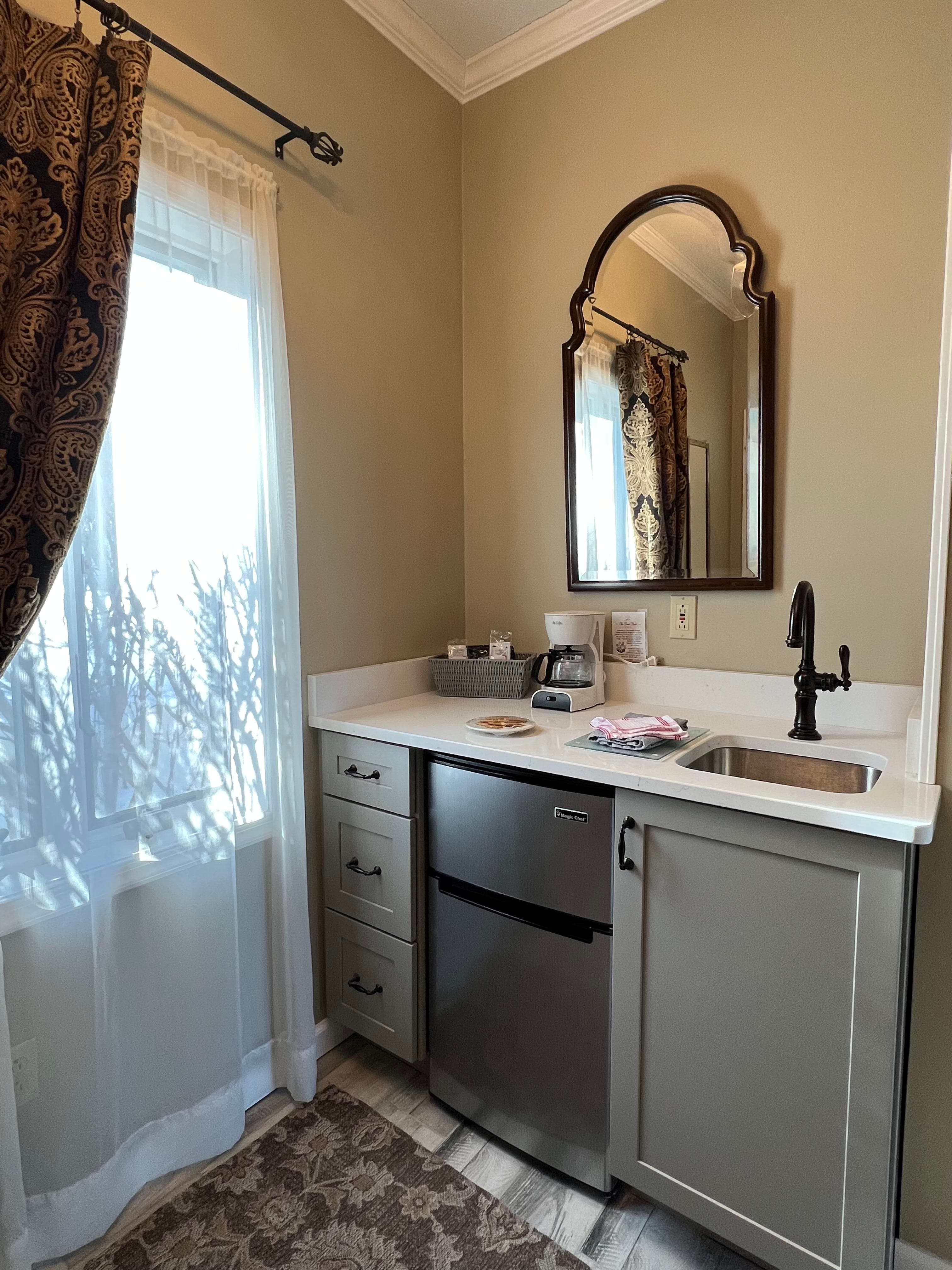 A small kitchen nook with a sink, fridge, and mirror, featuring a window with sheer curtains.