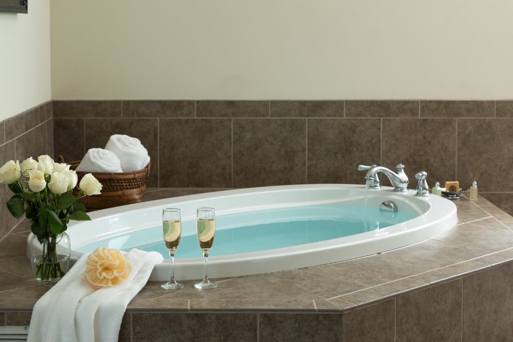 A luxurious oval bathtub filled with water, with two champagne flutes and a bouquet of white roses nearby.