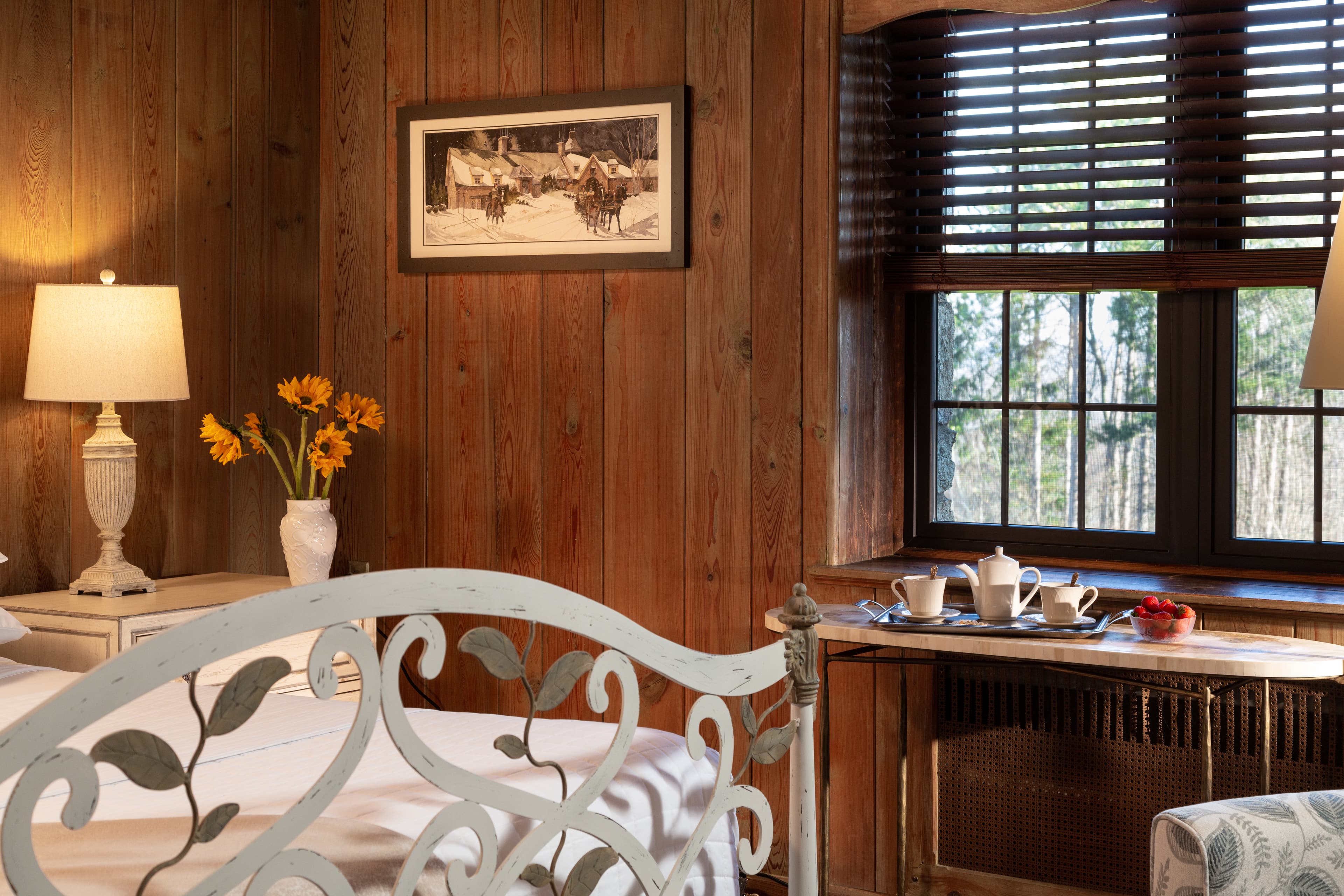 A cozy bedroom featuring a metal bed frame, wooden walls, a vintage photograph, and a sunny window nook with tea set and flowers.