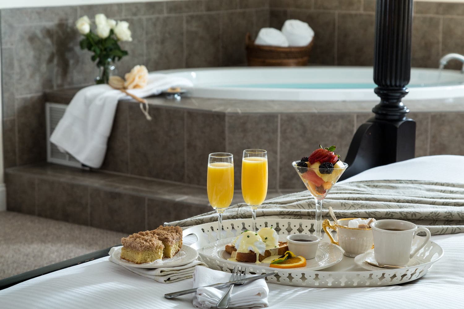 A luxurious hotel room featuring a tray of breakfast treats, two mimosas, and a jacuzzi in the background.