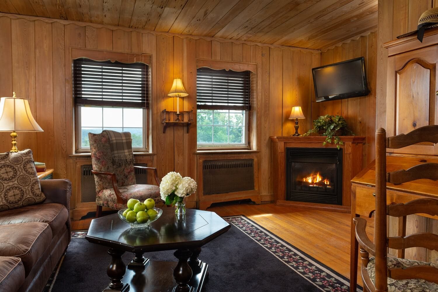 Cozy wooden-paneled living room with a fireplace, comfortable seating, and a coffee table displaying green apples and flowers.