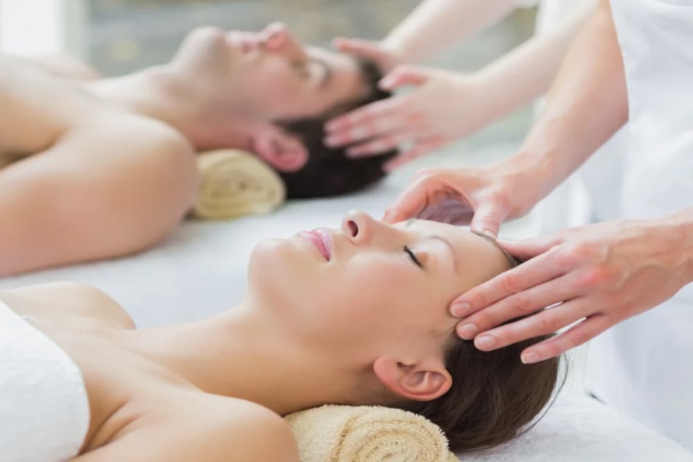 a man and woman laying on a massage table with a beige towel rolled under their necks, getting head massages.