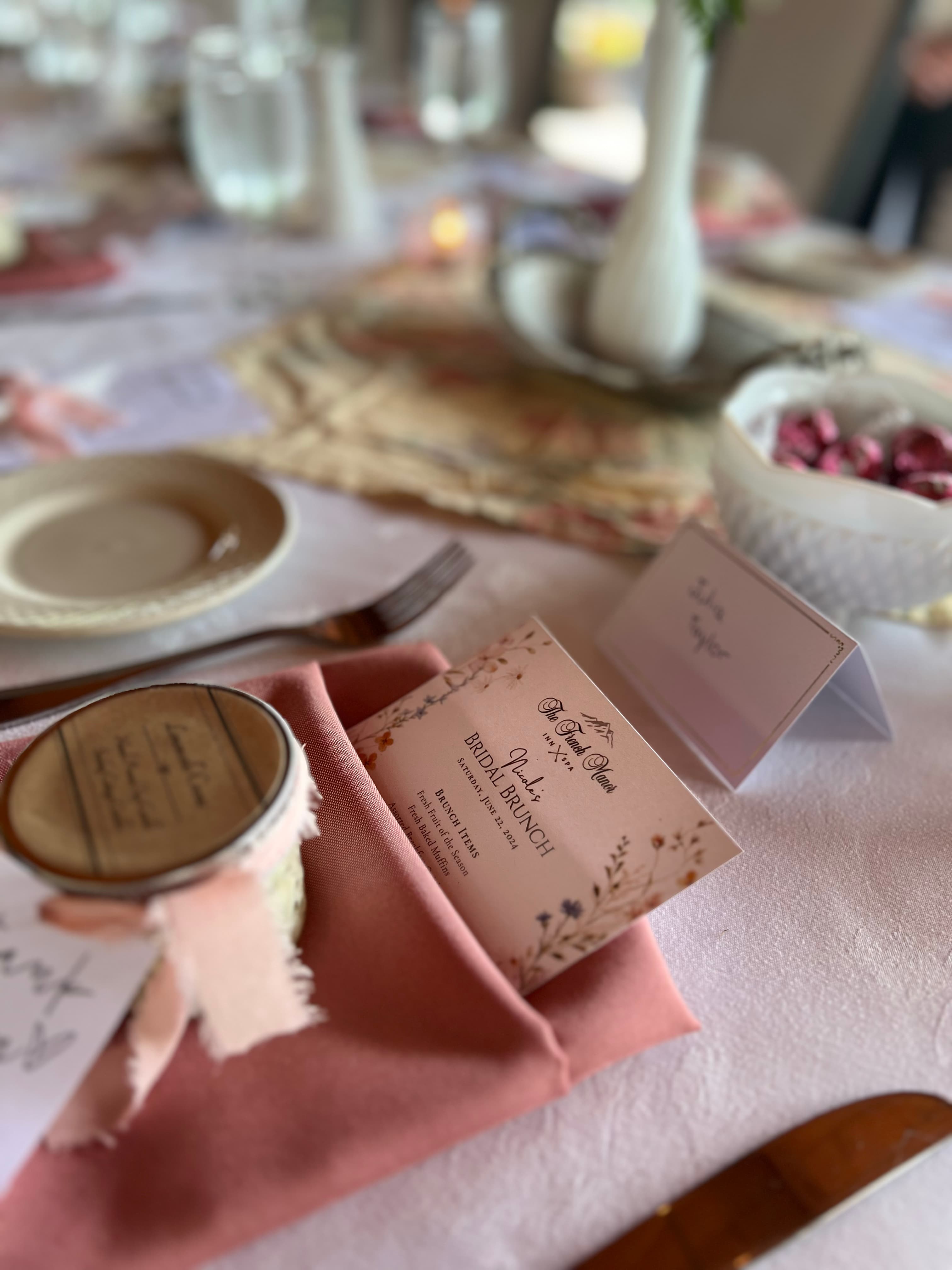A close-up of a place setting at a bridal brunch, featuring a peach-colored napkin, a personalized favor, a place card, and a glass of water.