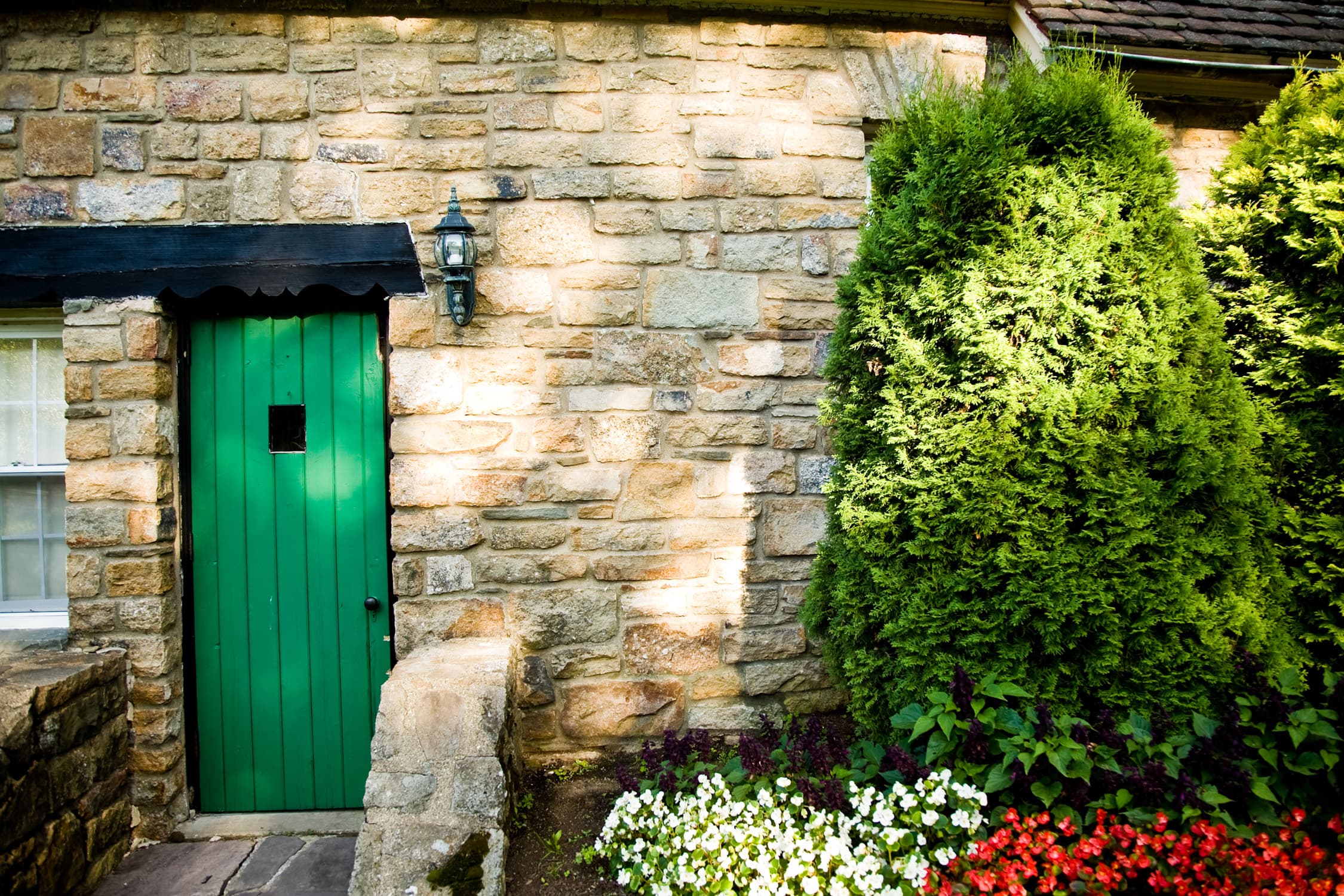 A green door with a small window is framed by stone walls and lush greenery.