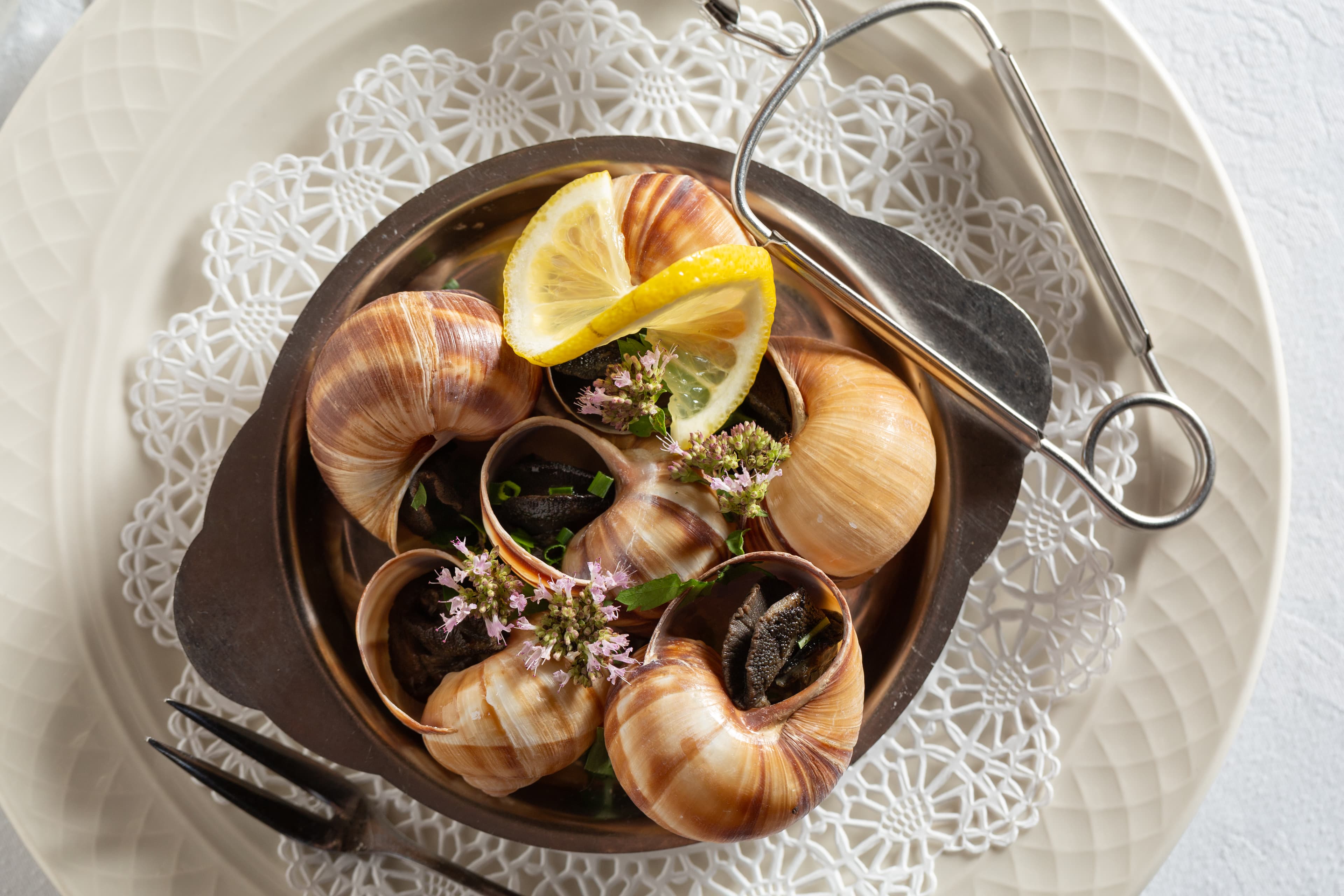 A dish of escargots de Bourgogne, featuring cooked snails served in their shells with a garlic butter sauce, herbs, and a lemon wedge. A special snail fork is also included on the plate.