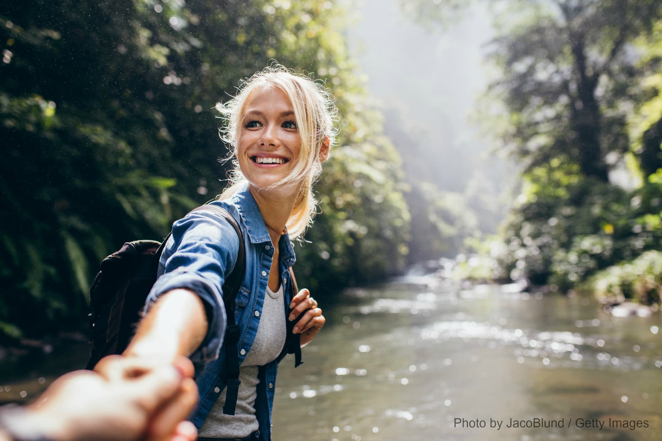 A smiling woman reaches out to hold hands while standing near a serene river surrounded by lush greenery.