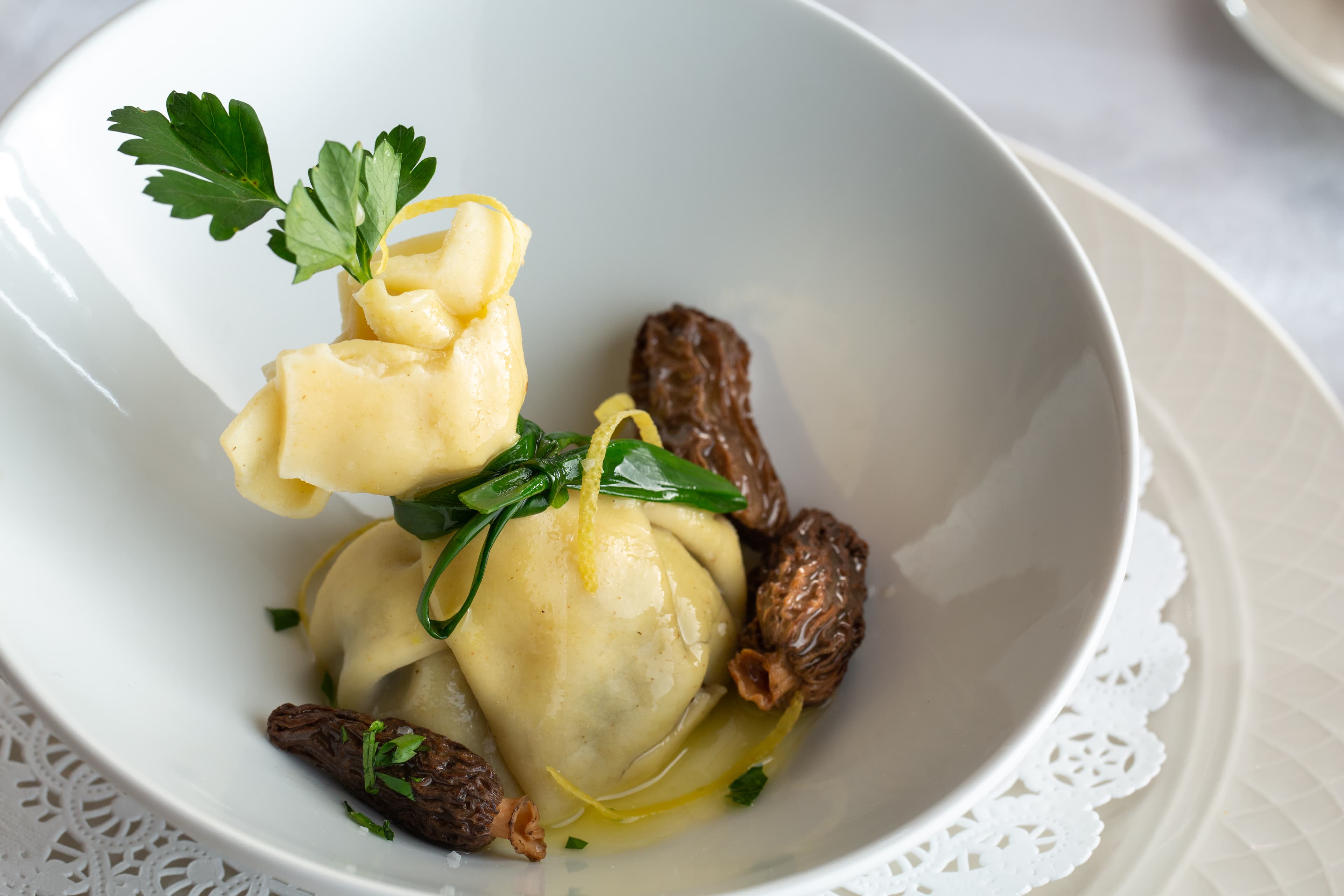 A delicate pasta dish called ravioli, shaped like a little purse tied with chives. The ravioli is filled with a creamy sauce and is served with sautéed morel mushrooms and a sprig of parsley.
