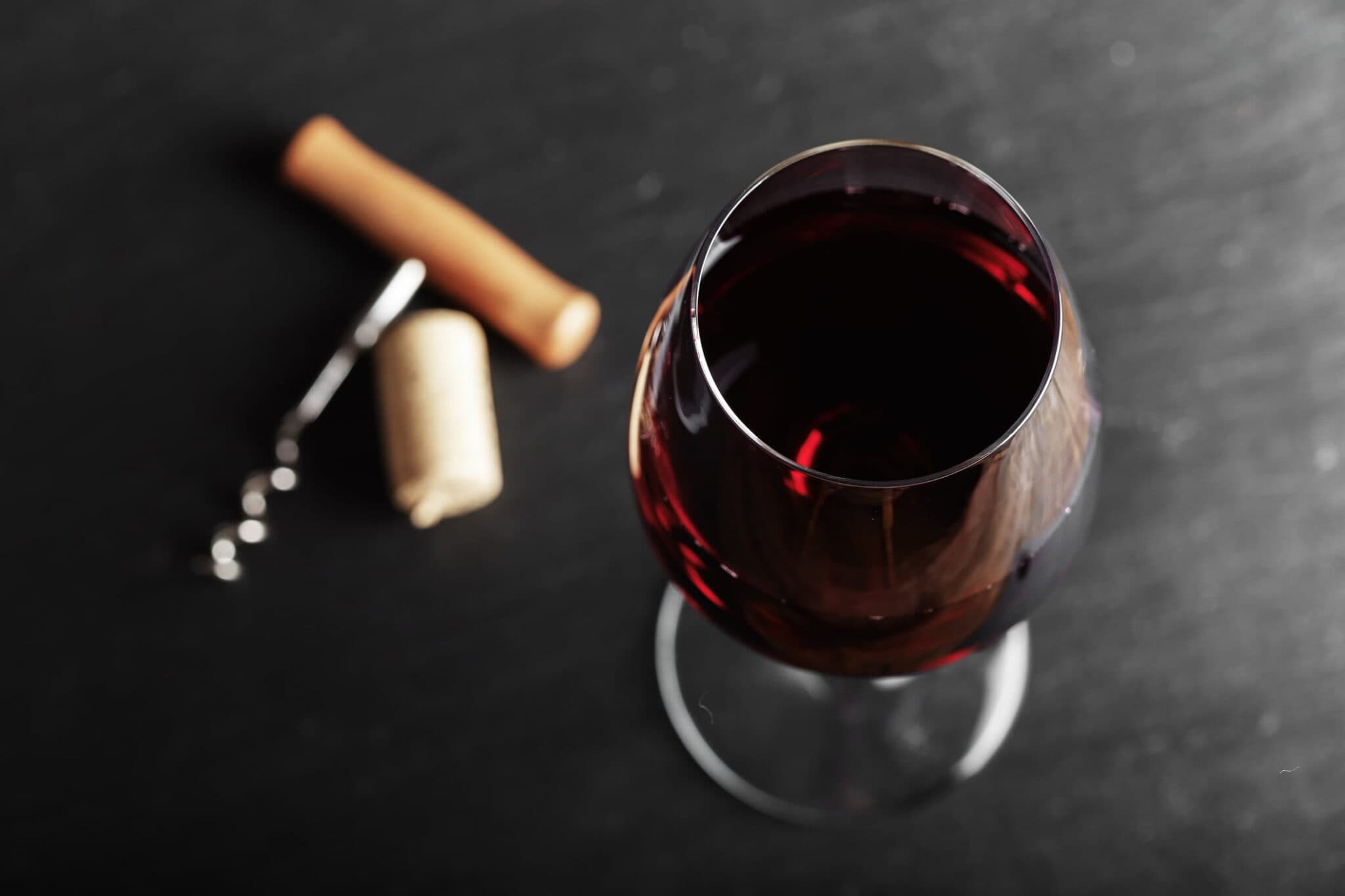 A glass of red wine with a cork and corkscrew beside it on a dark surface.