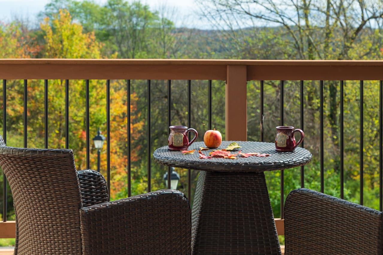 A cozy outdoor setting with a round wicker table, two mugs, an apple, and scattered leaves against a backdrop of colorful autumn trees.