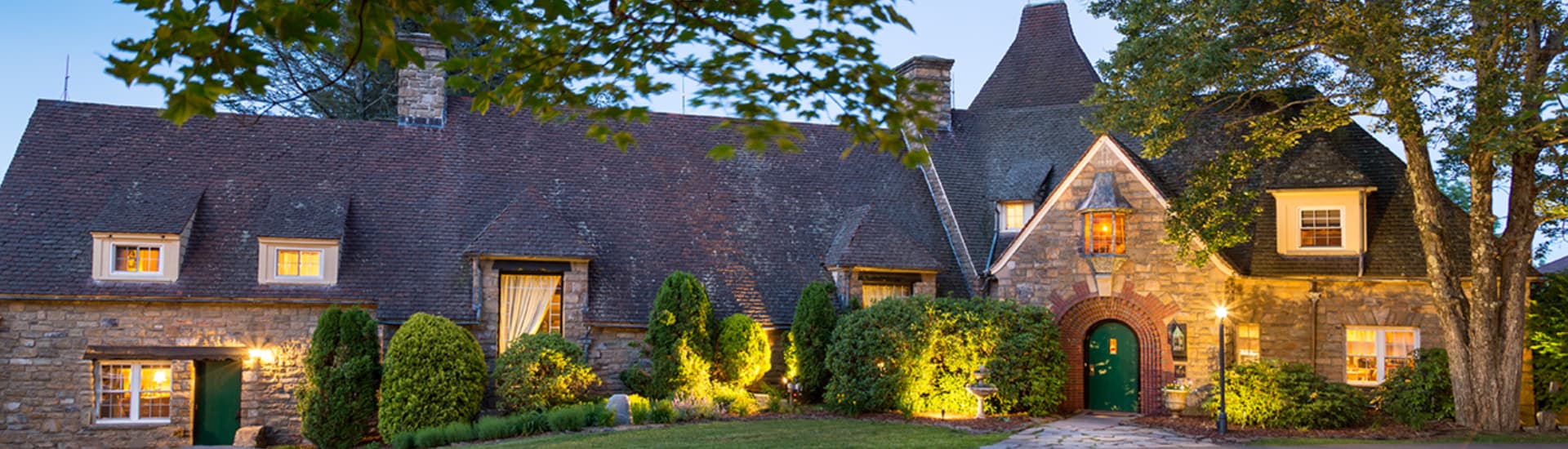 A charming stone mansion with a steeply pitched roof and a green front door. The mansion is surrounded by trees and shrubs, and warm lighting illuminates the facade at dusk.
