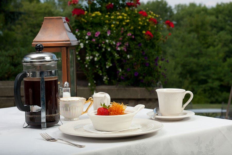 A table setting outdoors with a white plate holding a bowl of fruit and a cup of coffee. A French press coffee maker, a sugar bowl, and a creamer are also on the table. In the background, there is a lush garden with colorful flowers and a lantern.